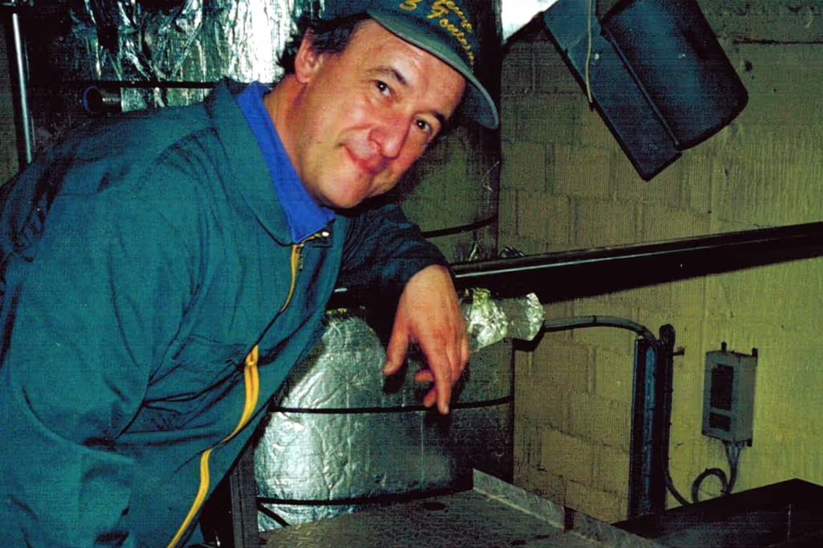 Man in a blue jacket and hat stands beside metal equipment in an industrial or kitchen setting, smiling at the camera.