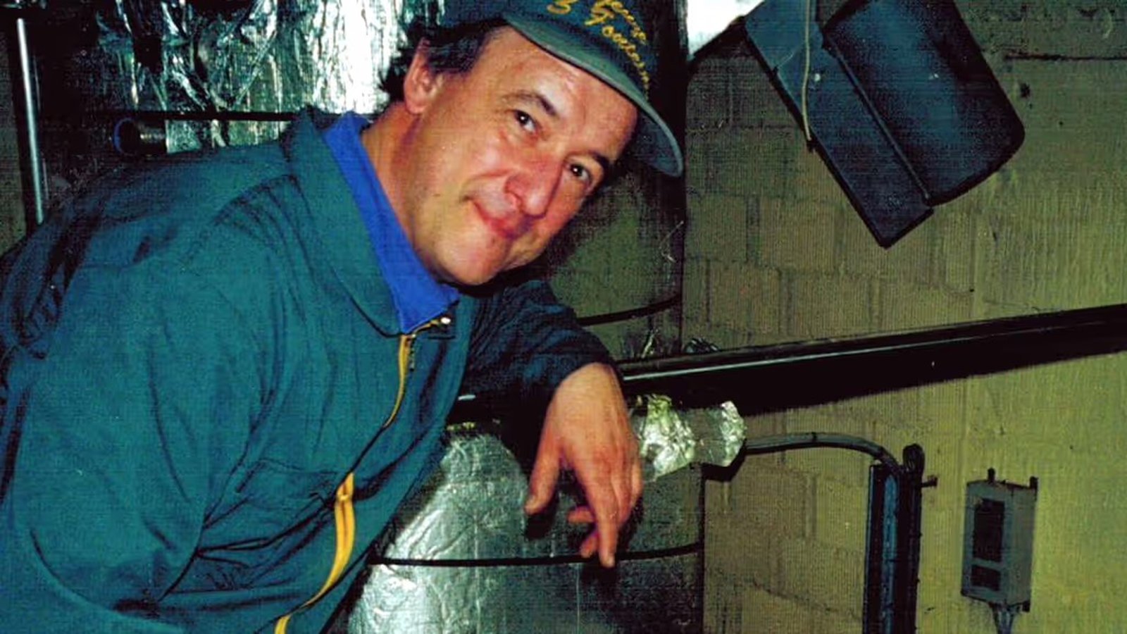Man in a blue jacket and hat stands beside metal equipment in an industrial or kitchen setting, smiling at the camera.