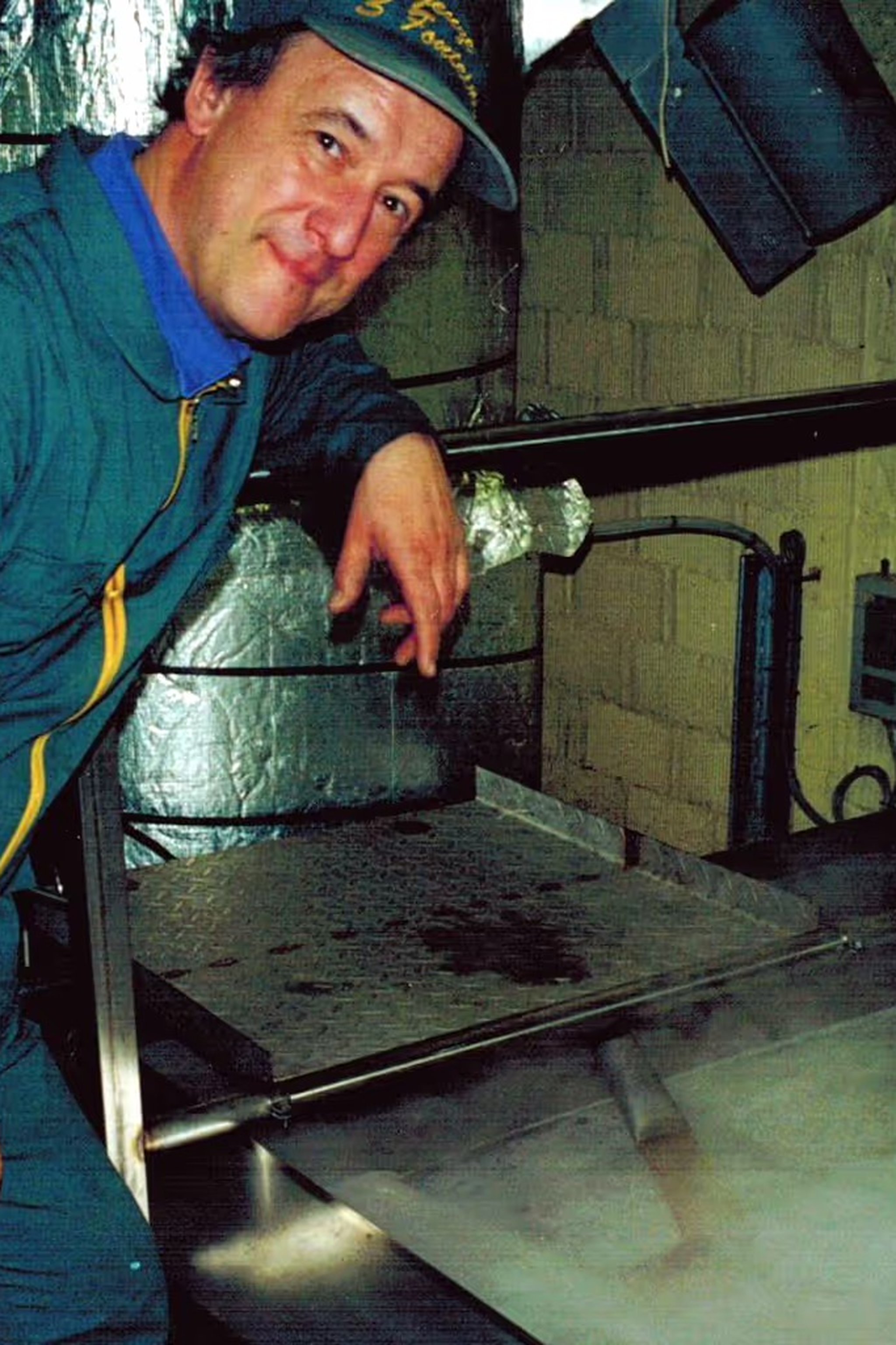 Man in a blue jacket and hat stands beside metal equipment in an industrial or kitchen setting, smiling at the camera.
