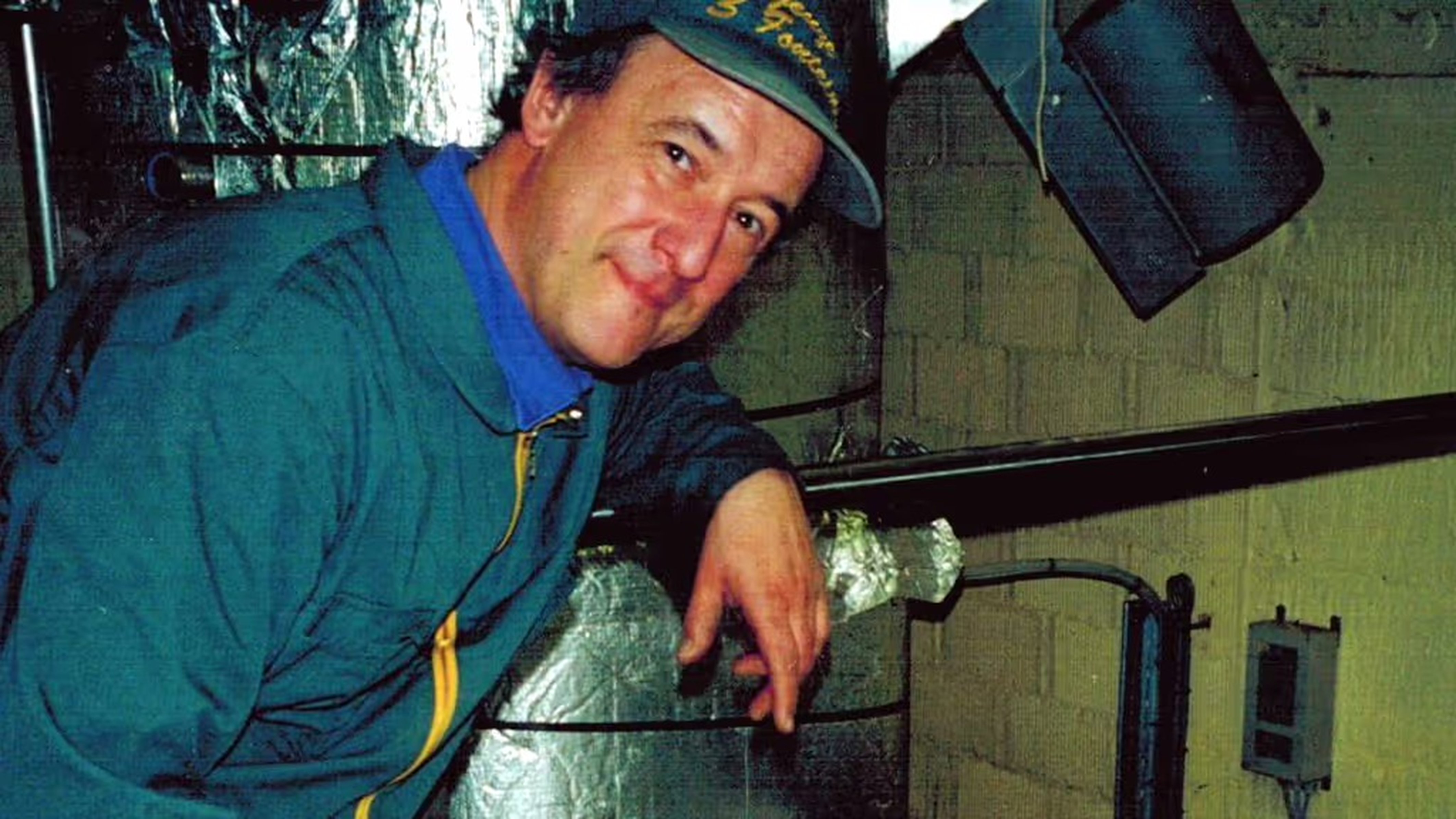Man in a blue jacket and hat stands beside metal equipment in an industrial or kitchen setting, smiling at the camera.