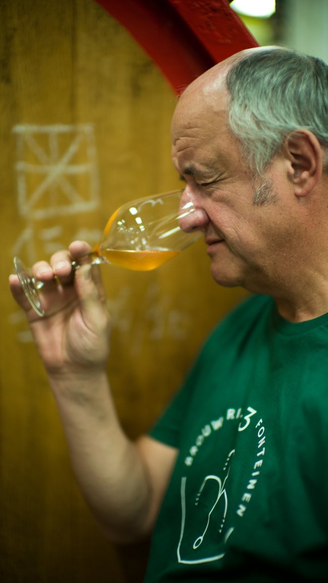 Armand wearing a green shirt, closely examining a glass of beer in front of wooden foeders.