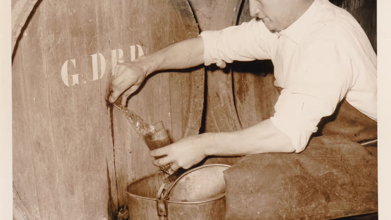 A black-and-white photo of Gaston Debelder crouching next to a wooden barrel, filling a jug with lambic.