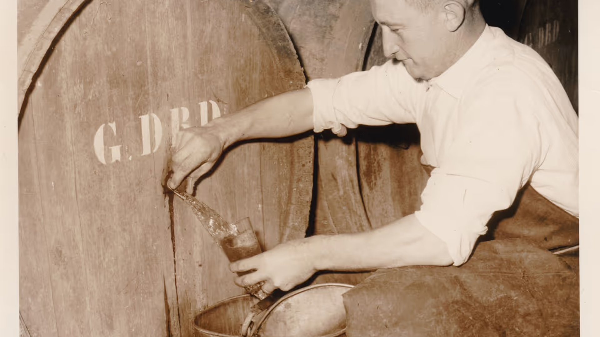A black-and-white photo of Gaston Debelder crouching next to a wooden barrel, filling a jug with lambic.