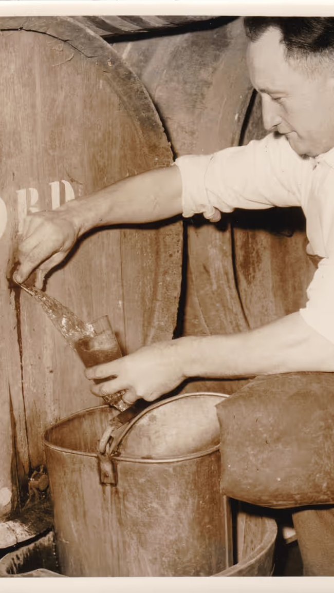 A black-and-white photo of Gaston Debelder crouching next to a wooden barrel, filling a jug with lambic.