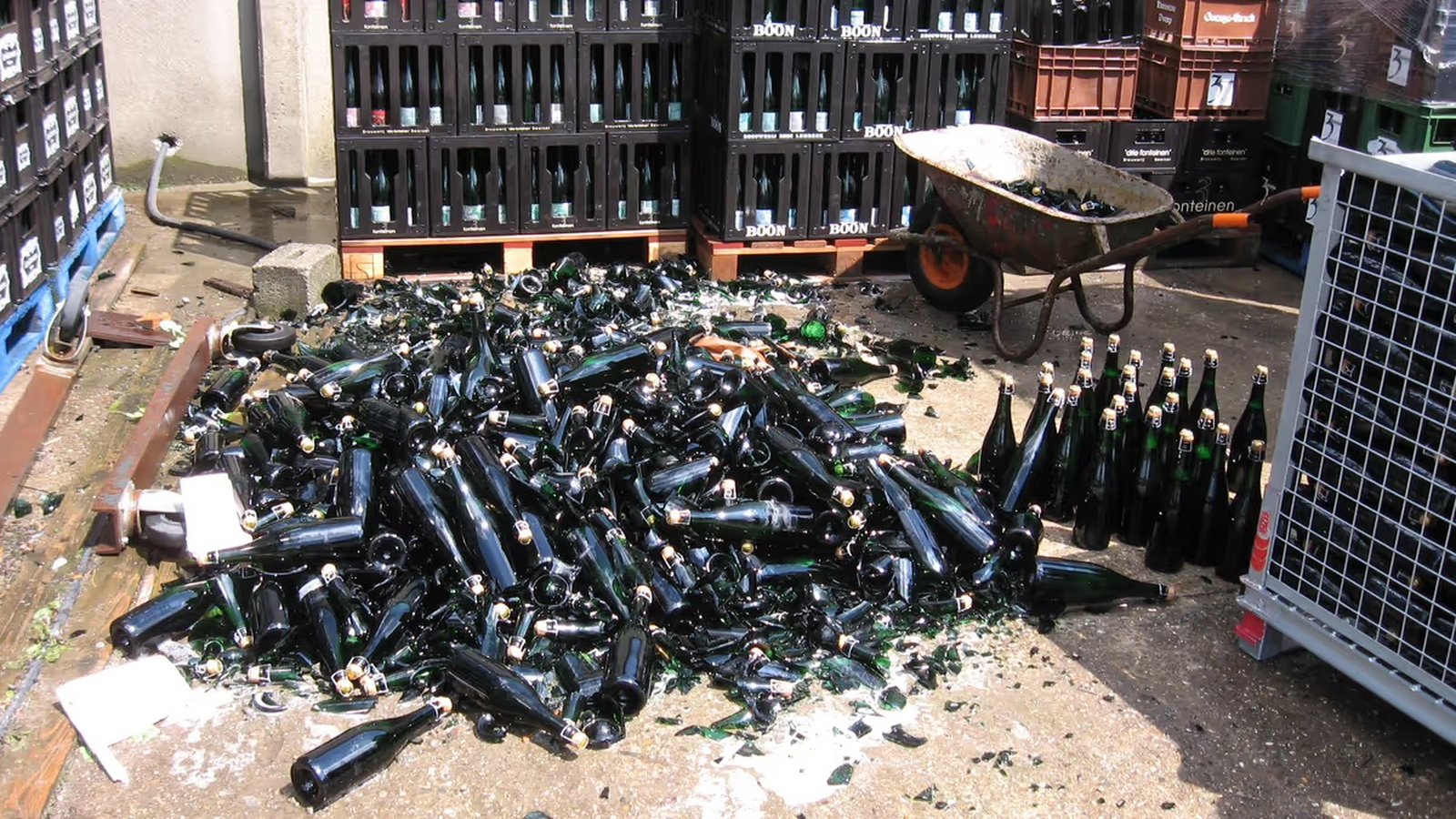 A pile of broken and intact green glass bottles lies on the ground next to crates and a wheelbarrow in an industrial area.