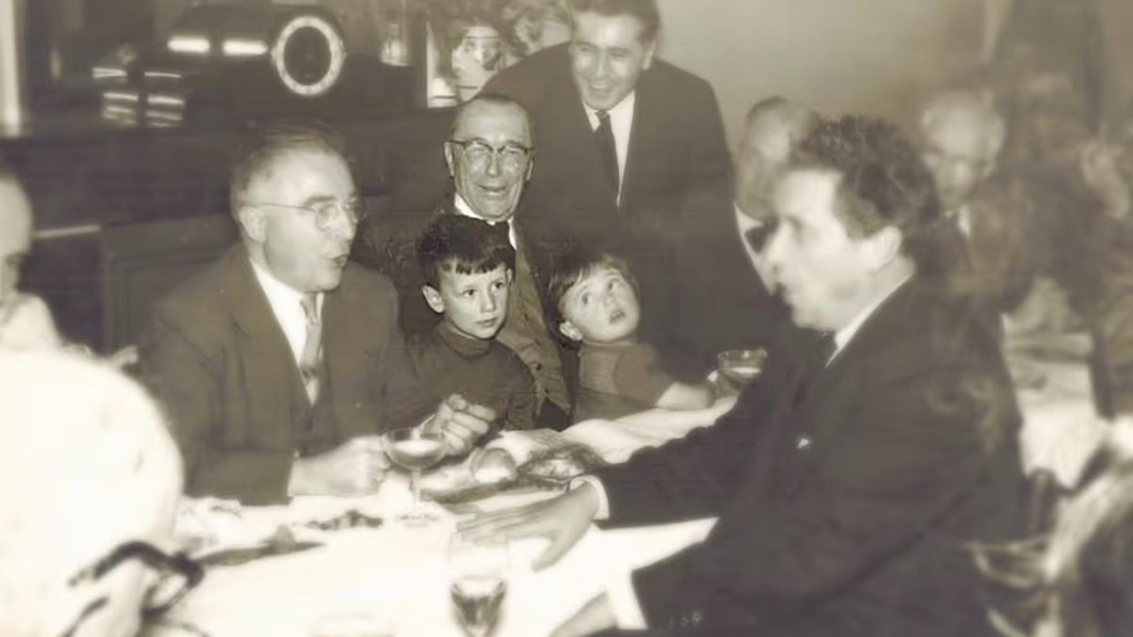 Vintage photo of people gathered around a dining table, with two children at the center, engaged in conversation.