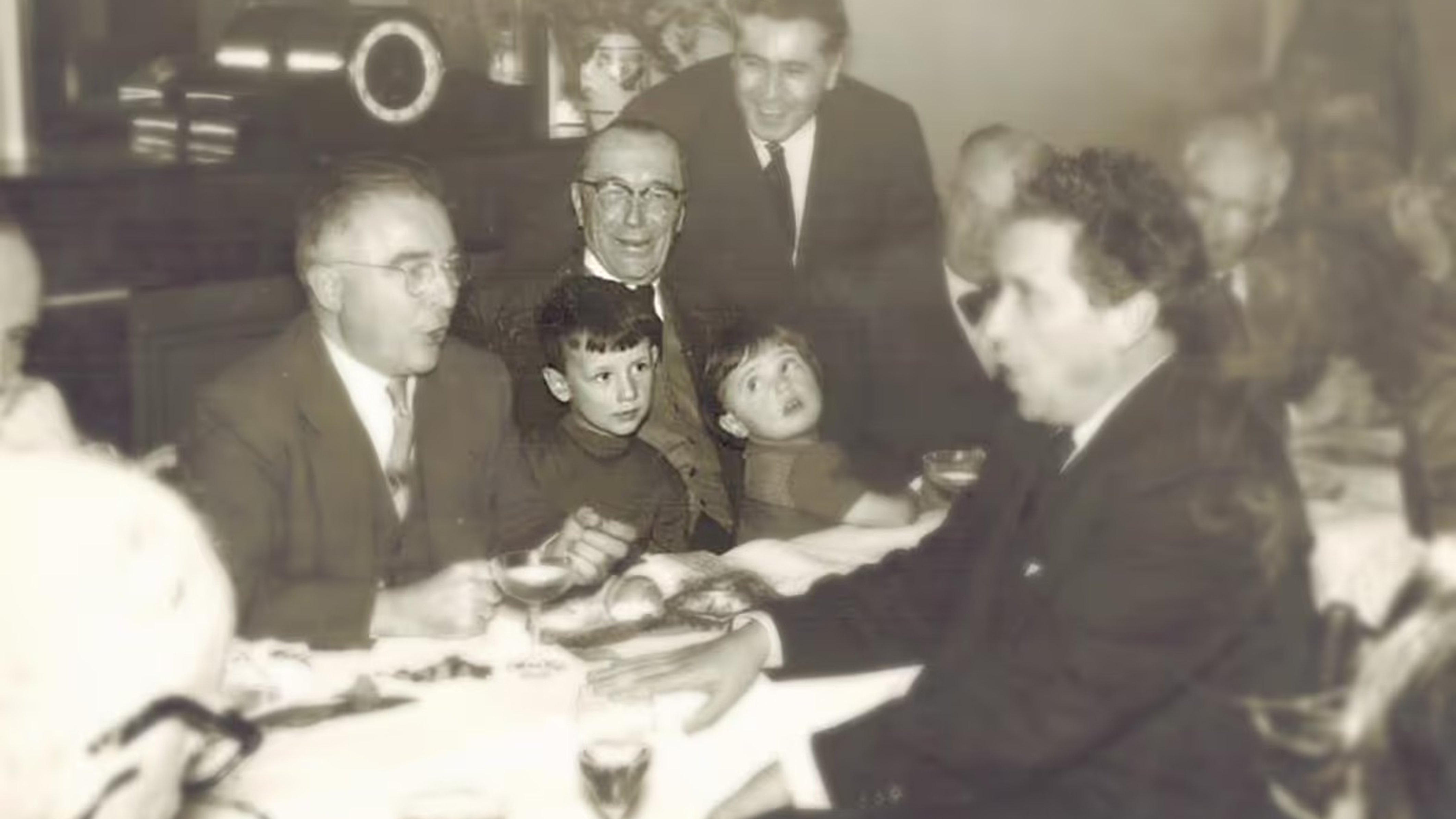 Vintage photo of people gathered around a dining table, with two children at the center, engaged in conversation.