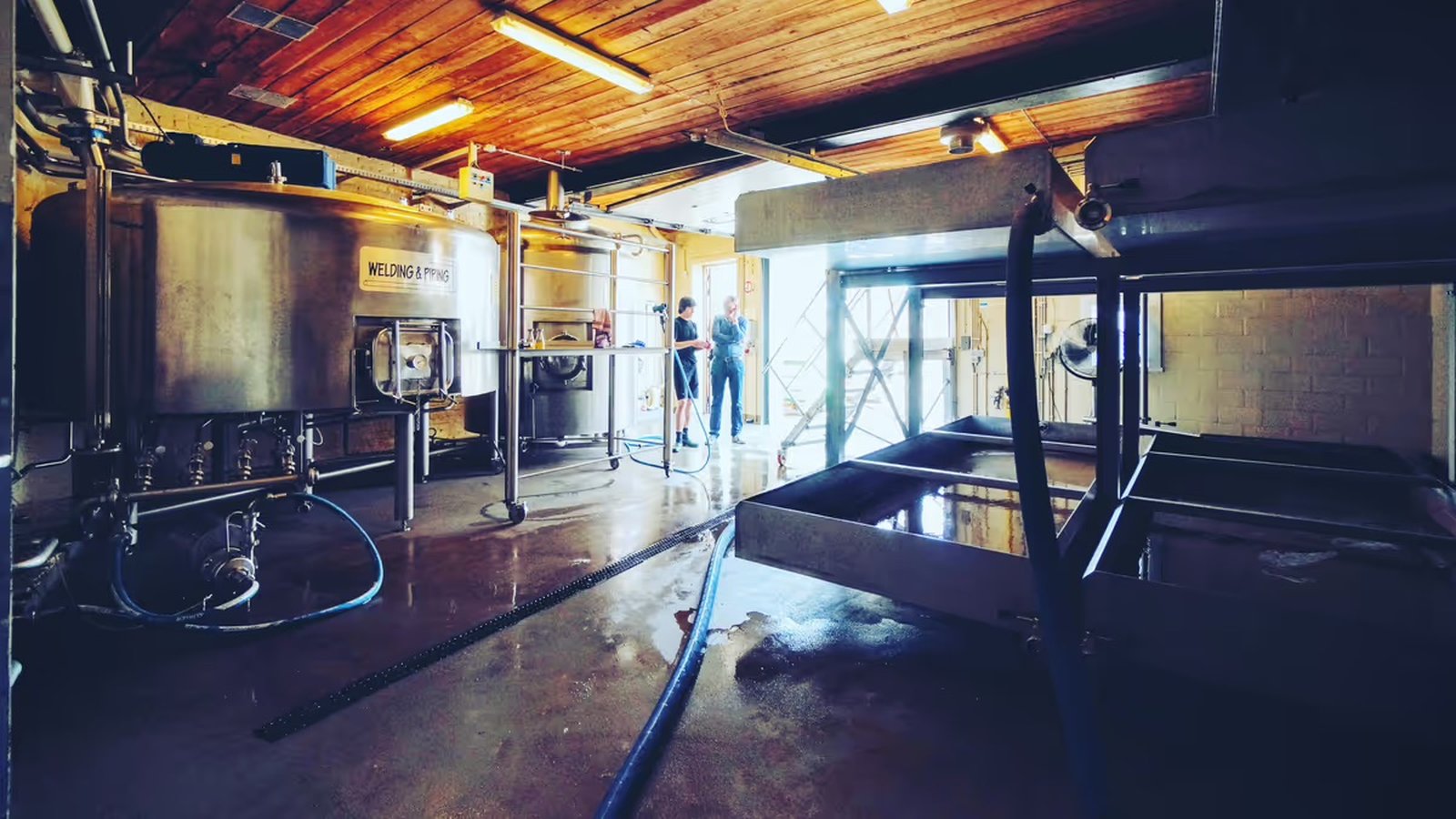 Interior of a brewery with large metal tanks, pipes, and equipment. Two people stand near the entrance, illuminated by natural light.