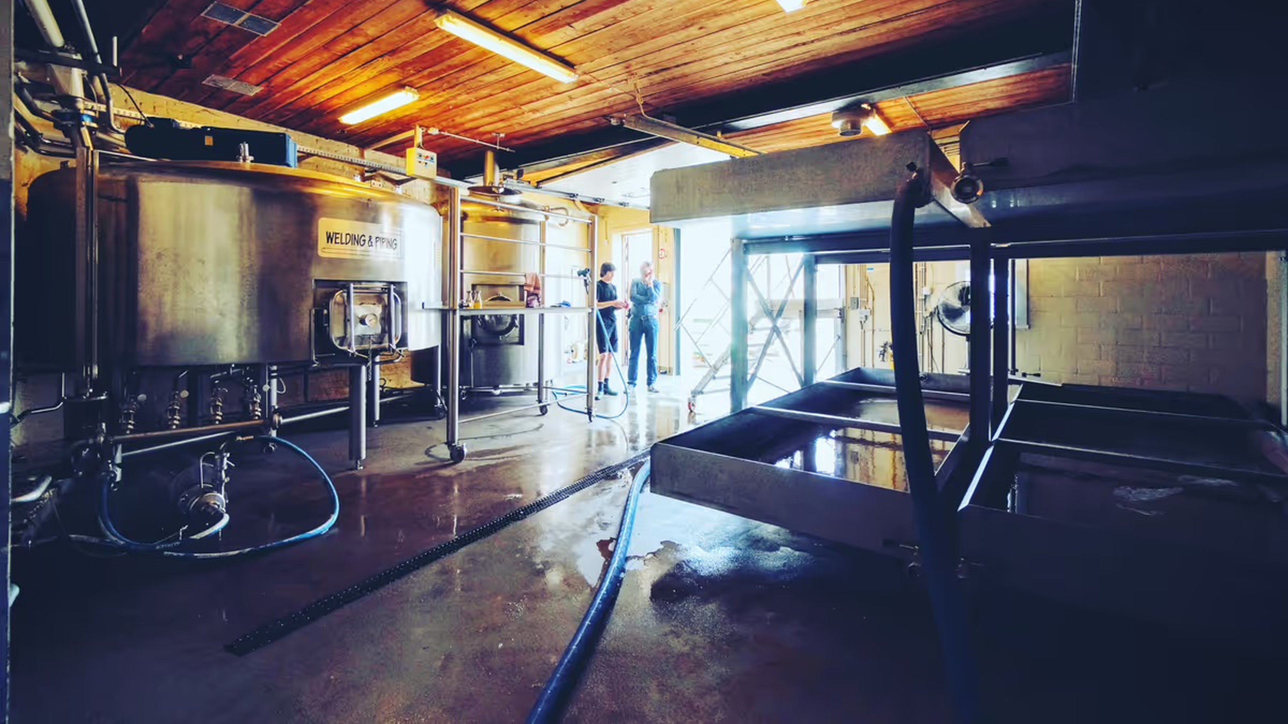 Interior of a brewery with large metal tanks, pipes, and equipment. Two people stand near the entrance, illuminated by natural light.