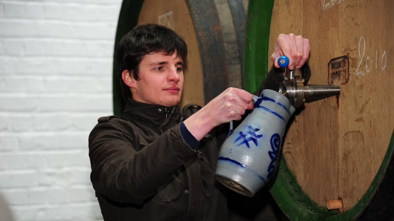 Person filling a ceramic jug with liquid from a large wooden barrel, wearing a dark jacket, in a setting with white brick walls.