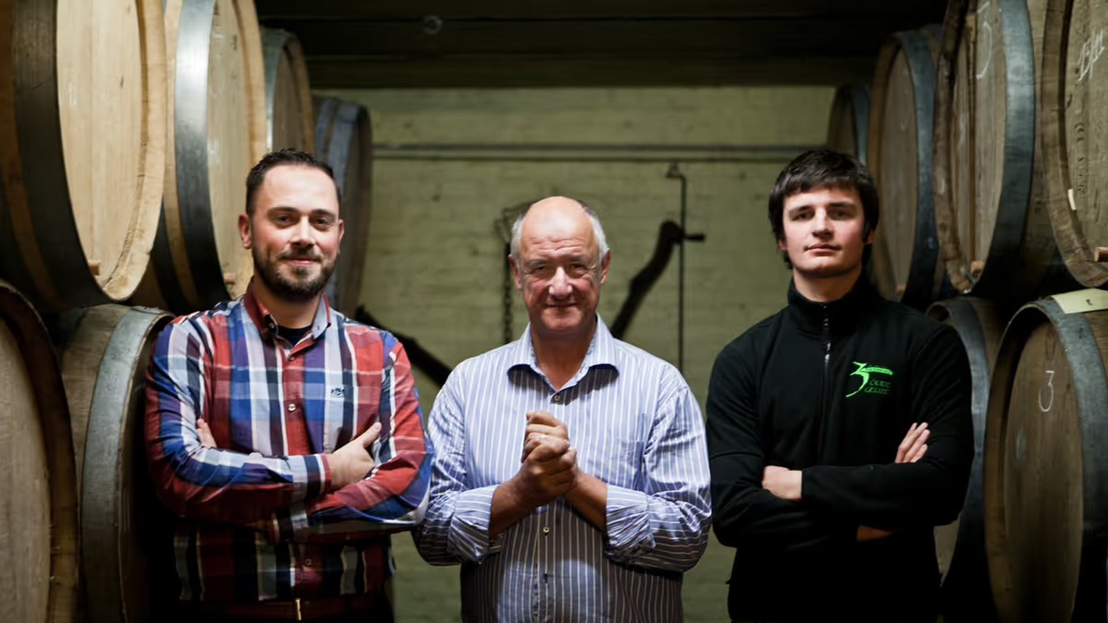 Armand Debelder standing proudly between Michaël Blancquaert and Werner Van Obberghen in the barrel room, smiling at the camera.