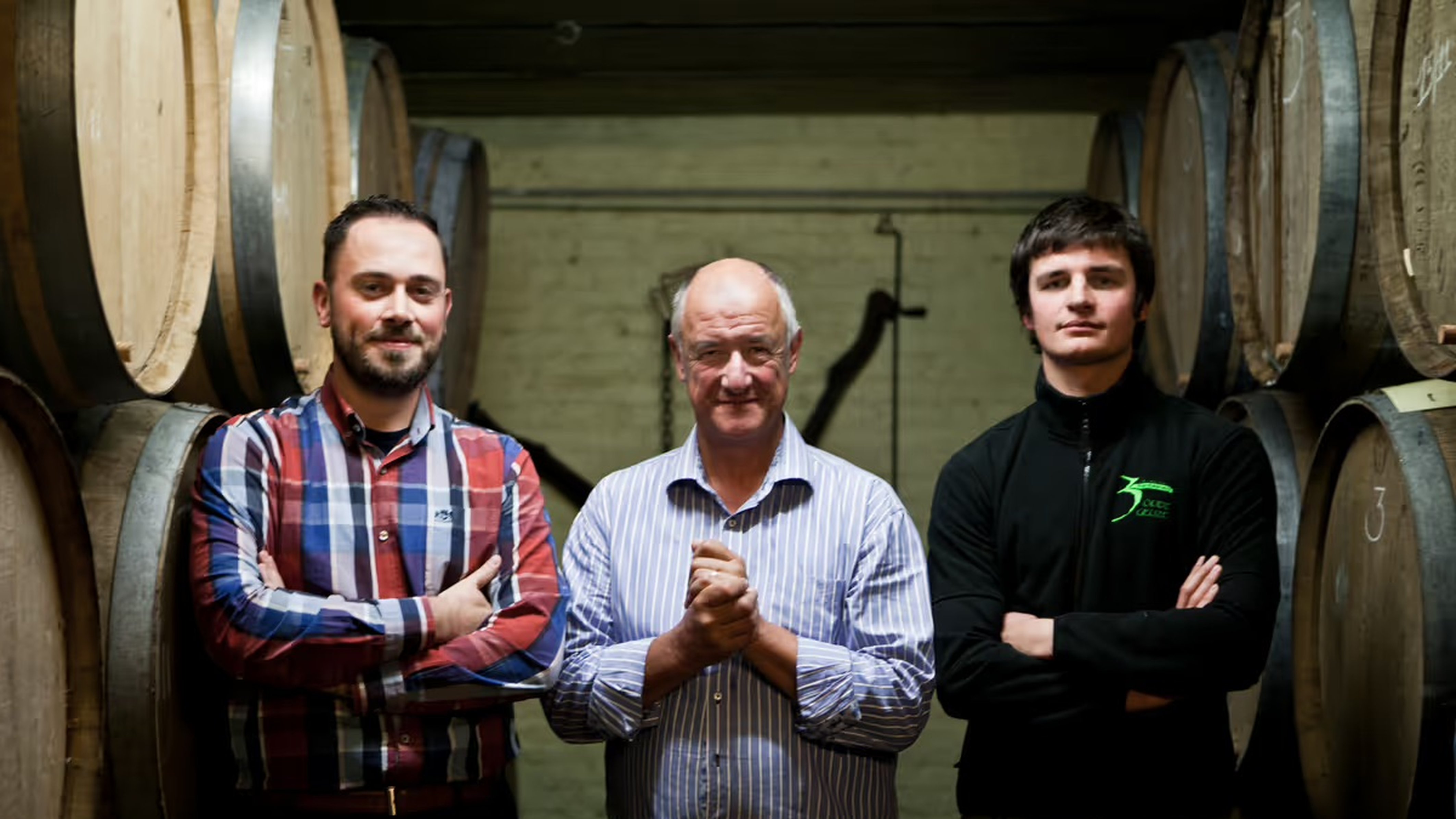 Armand Debelder standing proudly between Michaël Blancquaert and Werner Van Obberghen in the barrel room, smiling at the camera.