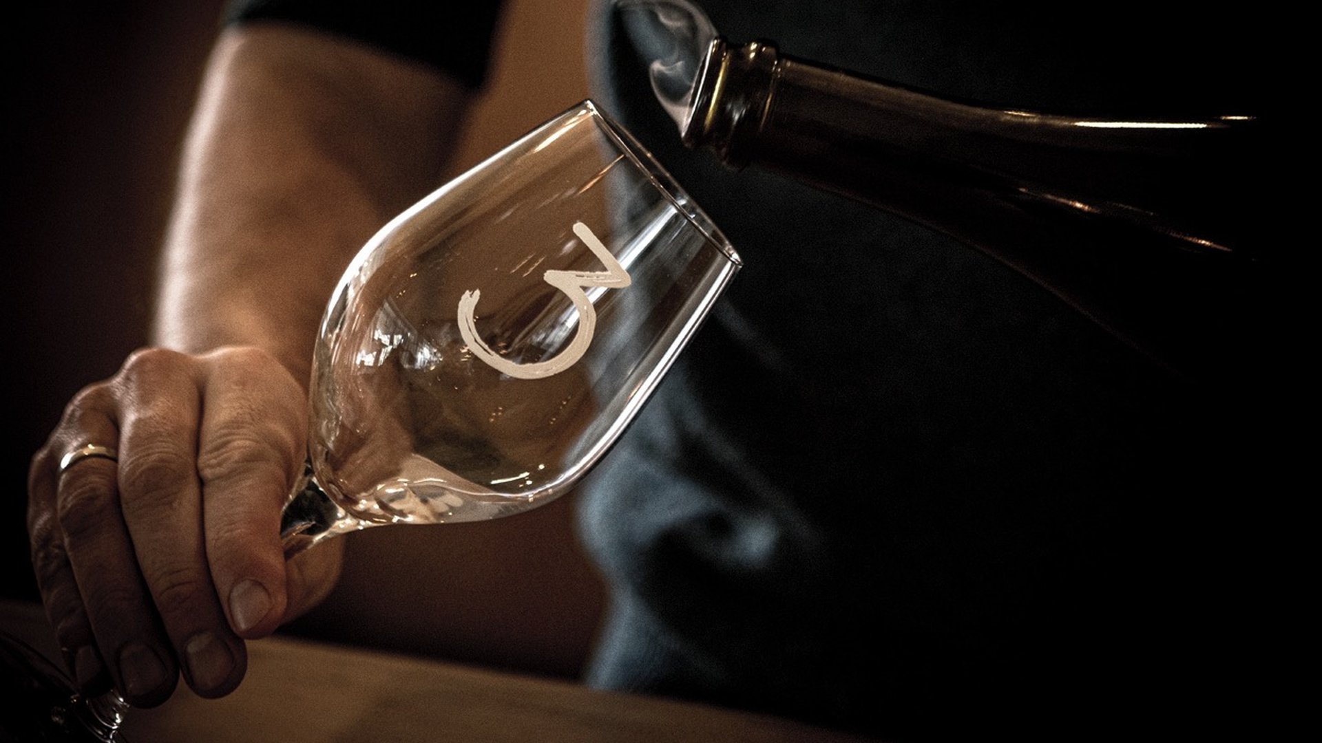 Close-up of a person pouring lambik into a tilted glass with a "C" logo, creating a sense of elegance and focus on the pouring action.