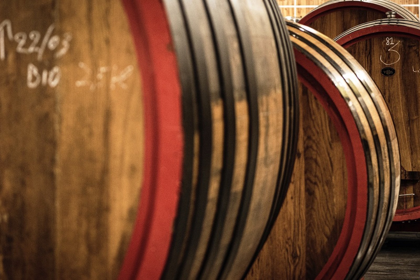 Close-up of large wooden barrels with red rims used for aging wine, with handwritten markings on the surface.
