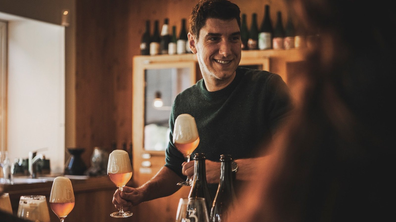 Man smiling while serving beer in wine glasses at a bar with wooden decor. Bottles are displayed in the background.