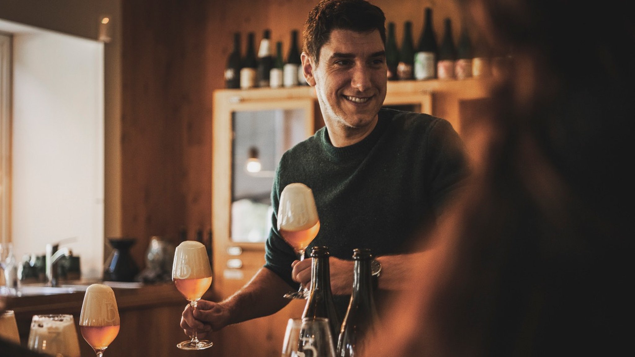 Man smiling while serving beer in wine glasses at a bar with wooden decor. Bottles are displayed in the background.