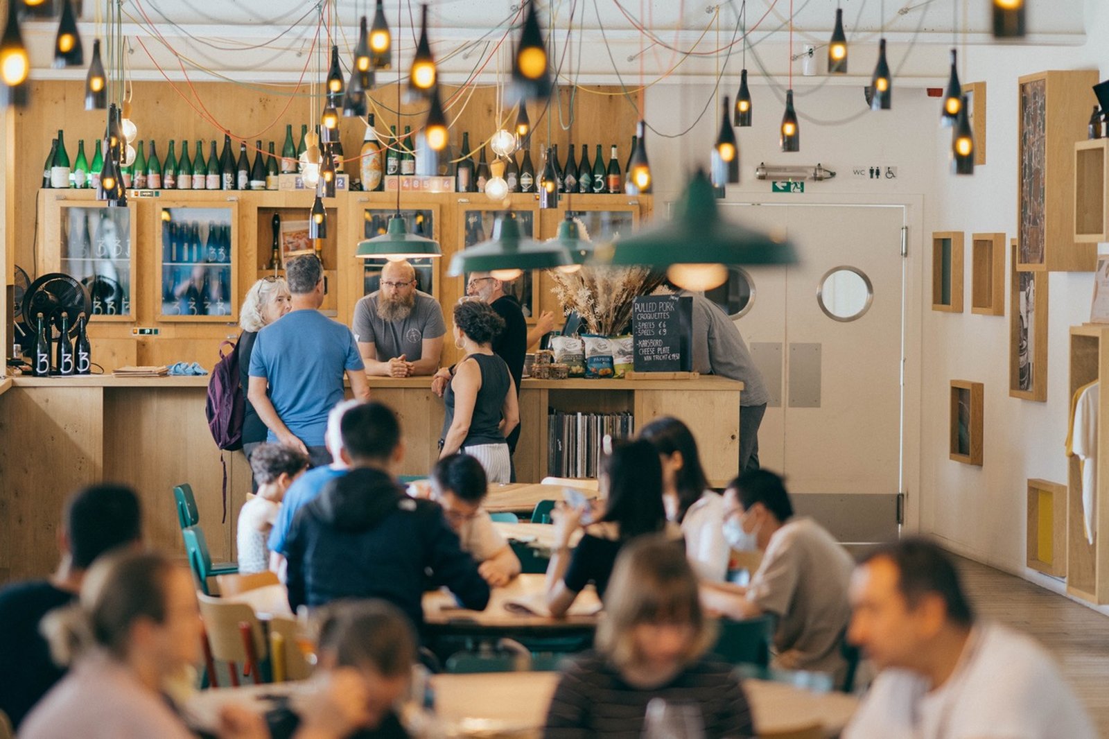 A cozy café with people gathered at the counter and seated at tables, warm lighting from hanging bulbs, and wooden accents throughout.