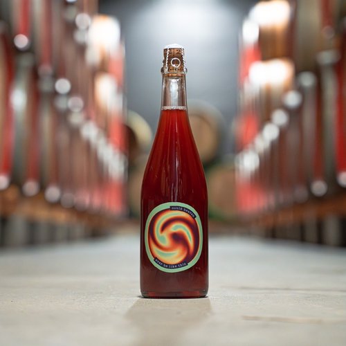 A bottle of the 3 Fonteinen and De Mederie collaboration standing in the middle of a barrel room aisle, flanked by rows of large oak foeders.