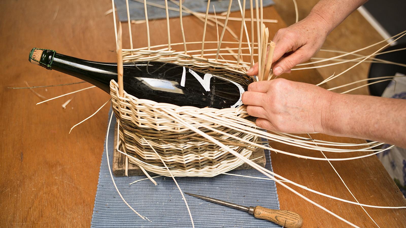 Hands weaving a wicker basket around a large bottle on a wooden table, with tools and reed strips scattered around.