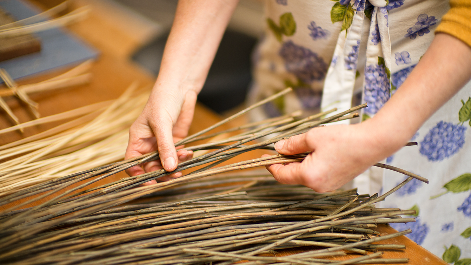 Person weaving with thin, long sticks on a table, wearing a floral apron and a yellow sweater.