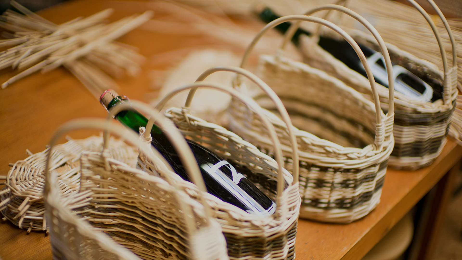Hands weaving a wicker basket around a large bottle on a wooden table, with tools and reed strips scattered around.