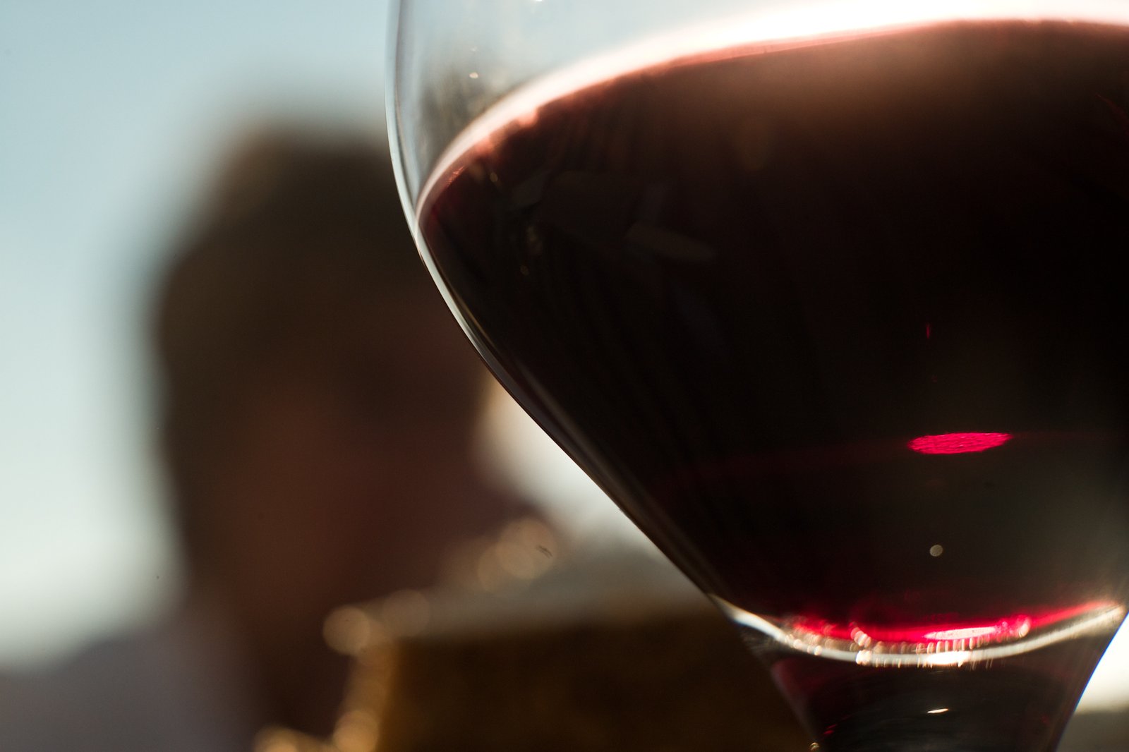 Close-up of a glass of dark red Oude Kriek, backlit by the sun, showing the deep colour and a silhouette of a person in the background.