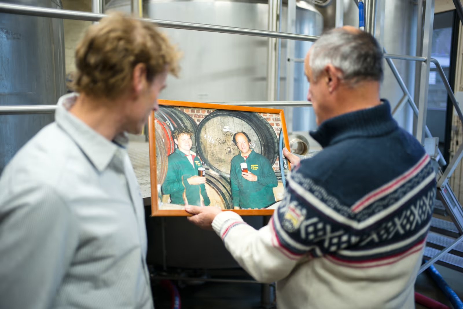 Two men in a brewery look at a framed photo of two people holding drinks, standing in front of large metal tanks.