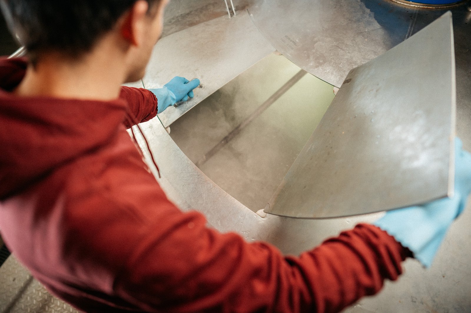 Person in red hoodie and blue gloves lifting a metal lid, revealing steam rising from inside a large industrial vat.
