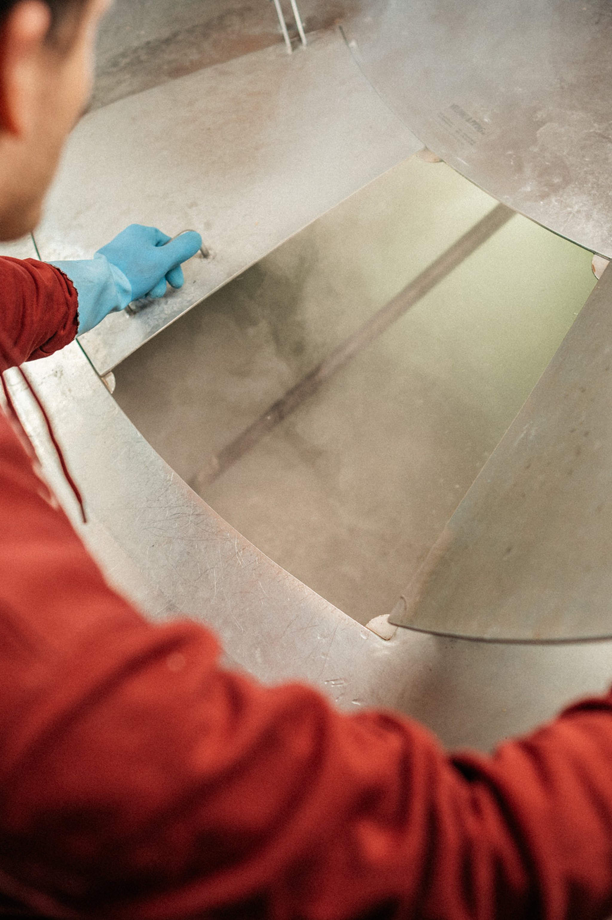 Person in red hoodie and blue gloves lifting a metal lid, revealing steam rising from inside a large industrial vat.