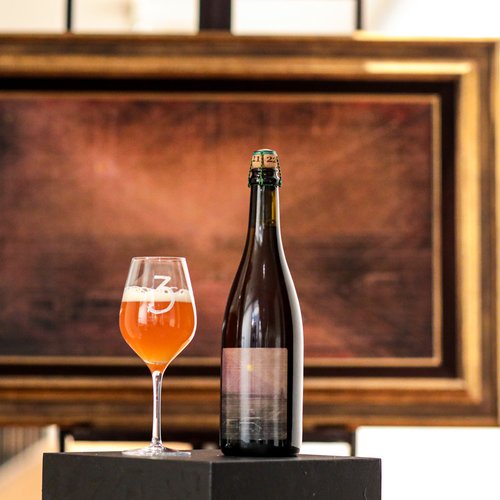 A 3 Fonteinen vintage bottle displayed on a black pedestal in front of a framed Felix De Boeck painting. The label on the bottle features a reproduction of the same artwork behind it.