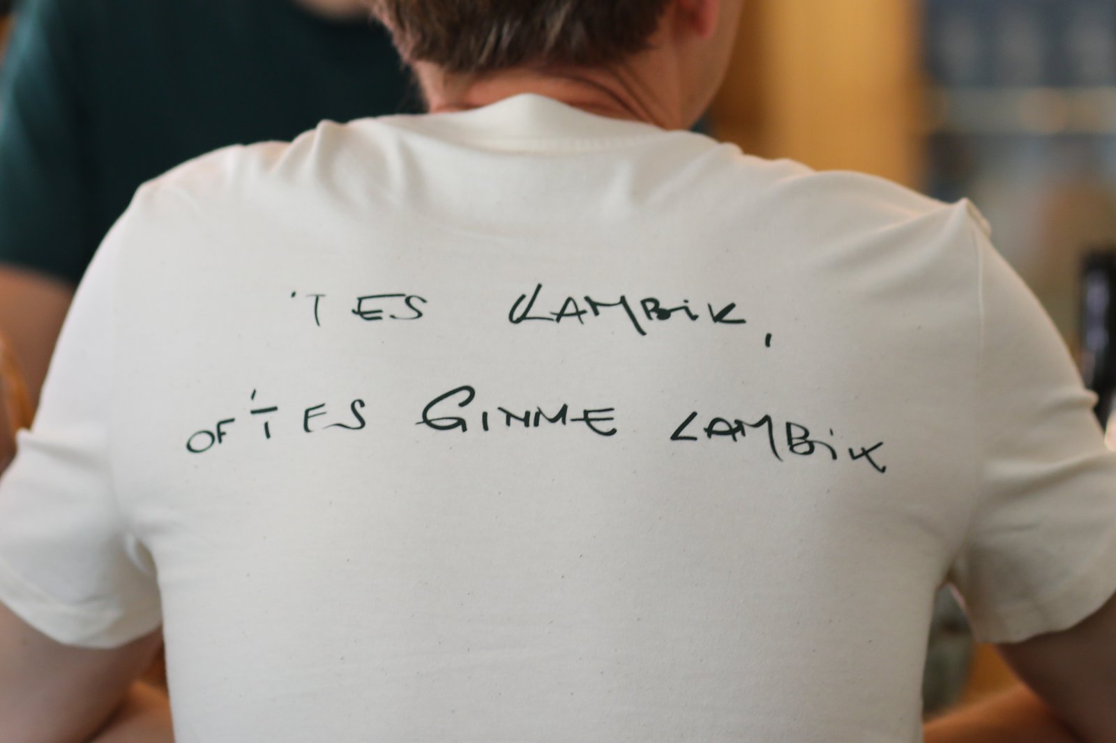 Person with short hair in a white t-shirt with black handwritten text at a bar, shelves with bottles in the background.