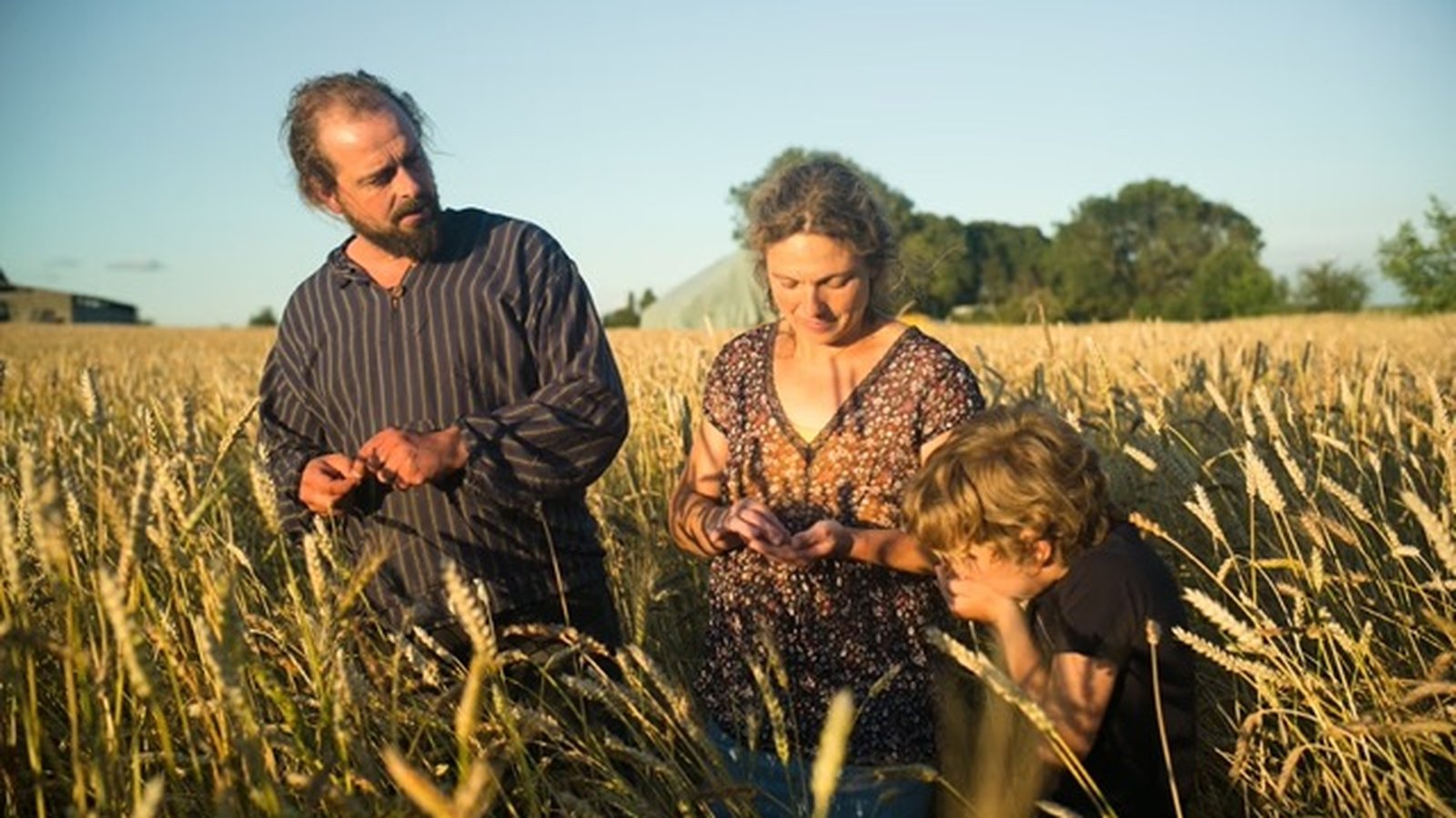 Farmers Hilde and Seppe standing together in a field, holding farming tools, with a barn structure in the background.