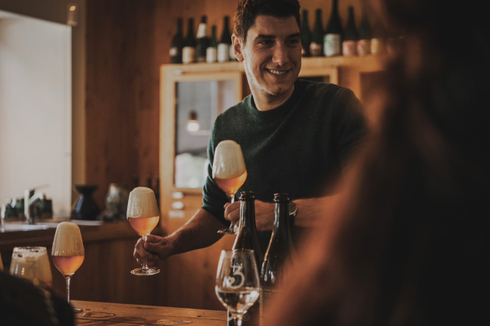 Man smiling while serving geuzes, standing at a wooden bar with bottles and a cabinet in the background.