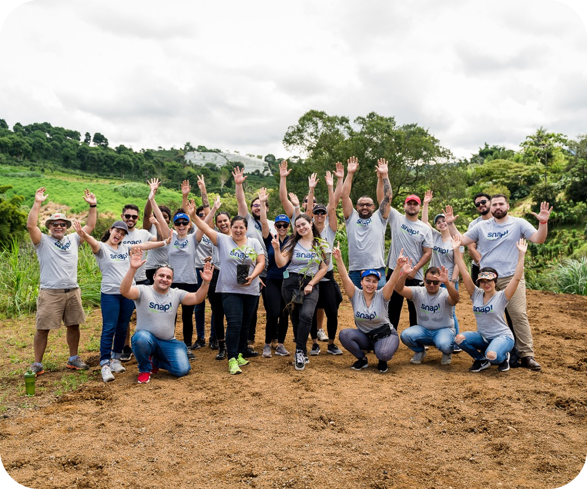 A group of smiling people in matching shirts raise their hands while standing on a dirt path with trees and greenery in the background.