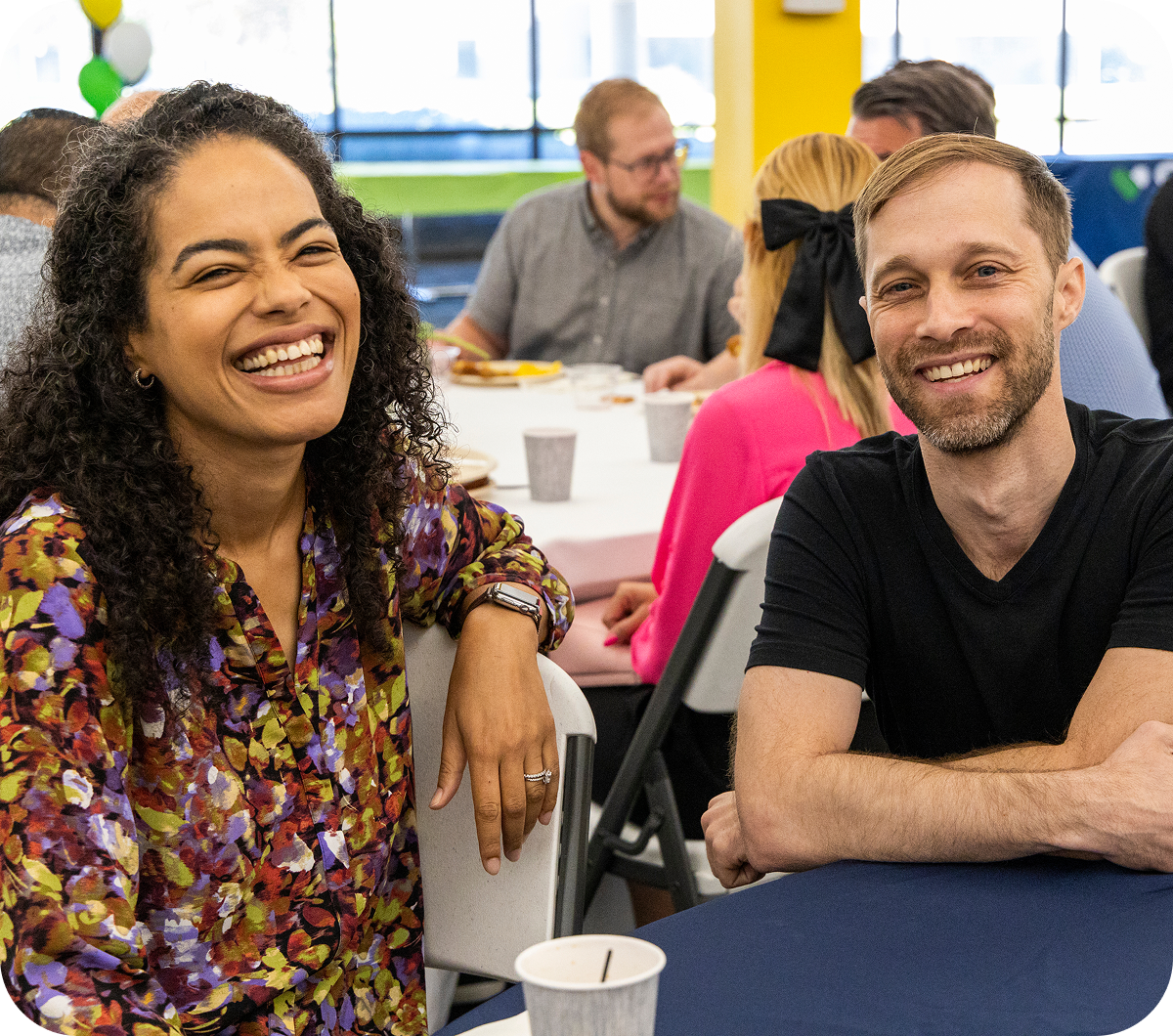 Two people smiling and sitting side by side at a table during a social event. Other attendees are visible in the background.