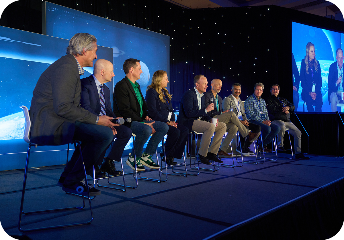 Nine people seated on a stage panel discussion, with a starry backdrop and a large screen displaying two of the panelists.