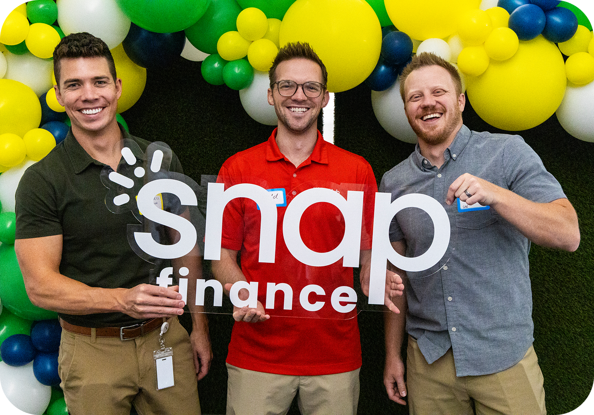 Three smiling men hold a "snap finance" sign in front of a balloon arch in green, yellow, blue, and white.