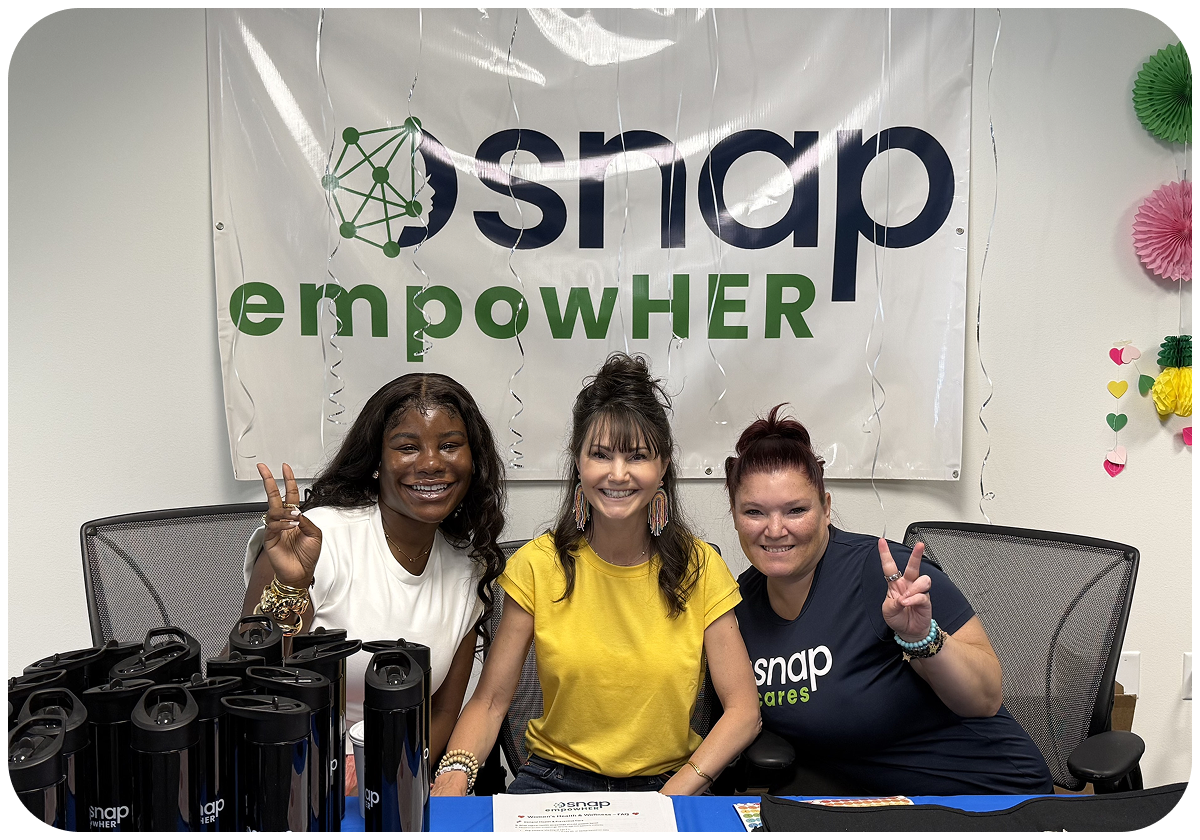 Three women smiling and making peace signs at a table with water bottles, in front of a "snap empowHER" banner.