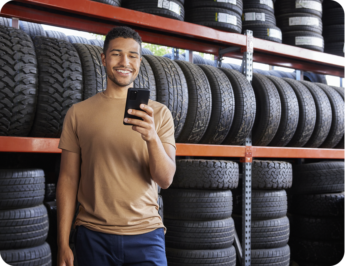 Man in a beige shirt smiles at his phone, standing in front of stacked tires on orange shelves in a store.