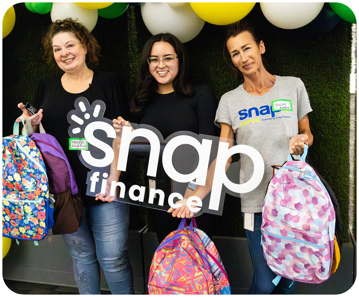 Three people holding colorful backpacks and a "snap finance" sign, standing in front of a wall with green and white balloons.