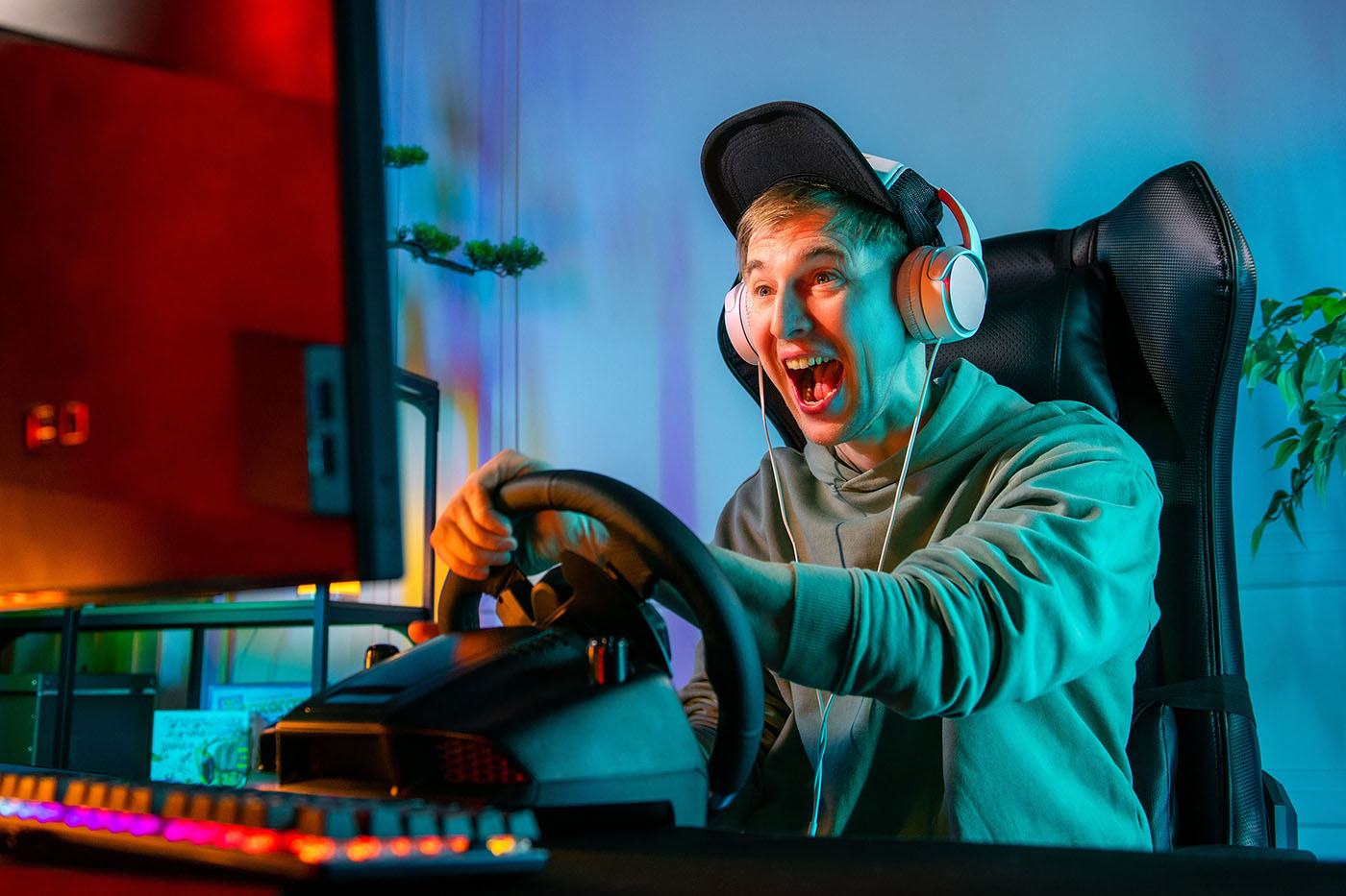 Excited gamer wearing headphones and cap grips a racing wheel at RGB-lit setup, shouting toward the monitor during a driving game.