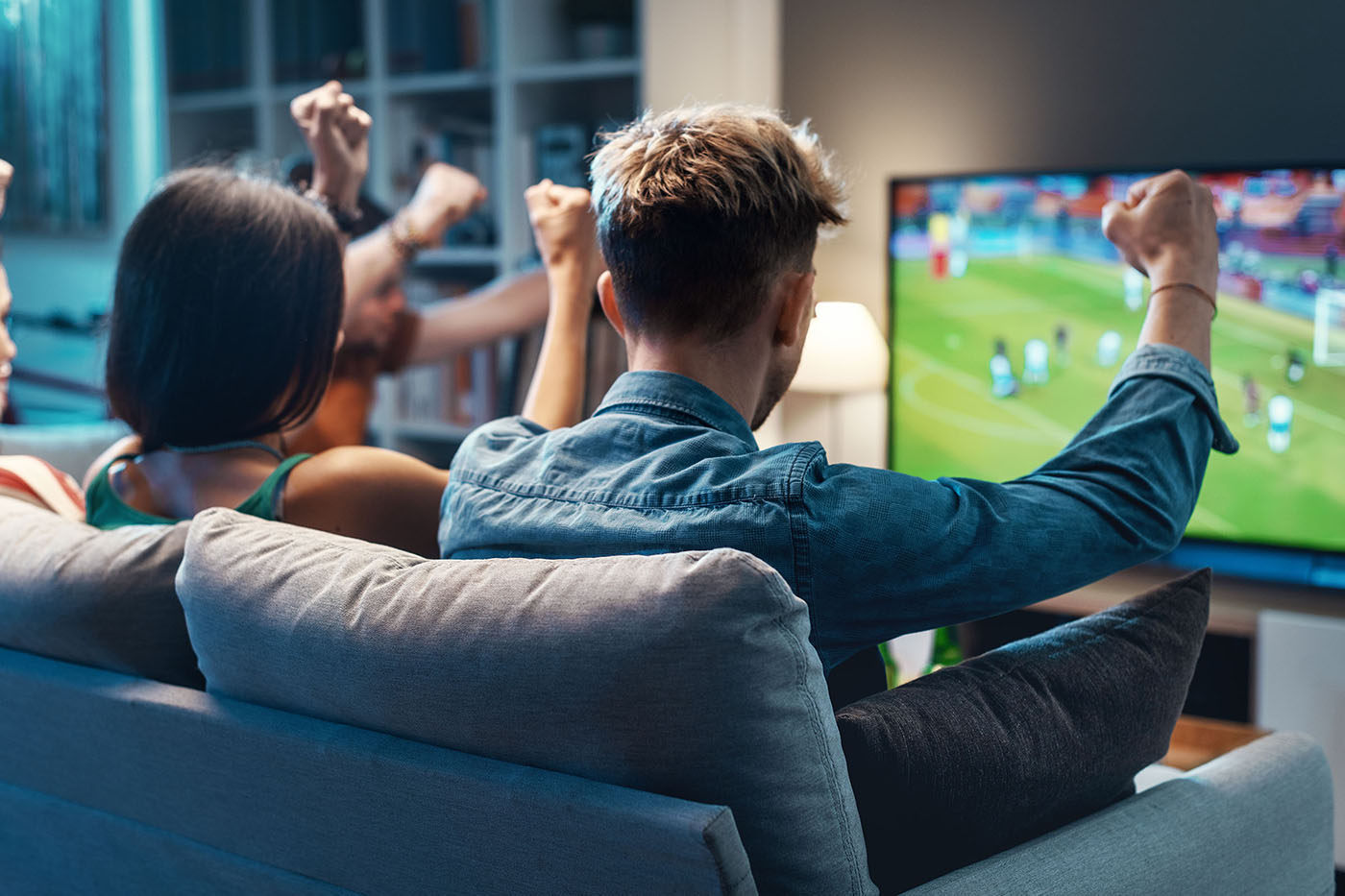 Friends on couch raise fists while watching soccer match on large TV in dimly lit living room.