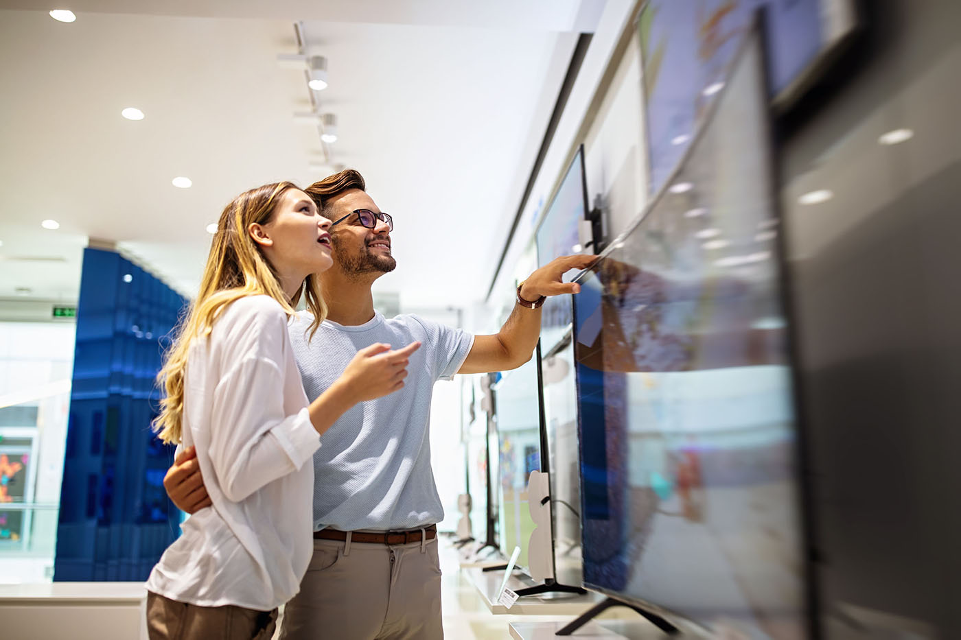 A couple stands in an electronics store, smiling and pointing at a large flat-screen TV on display.