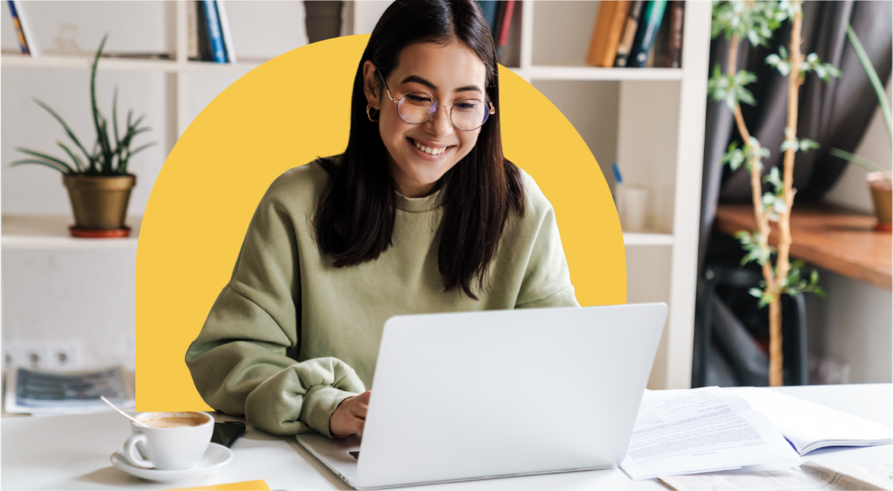 Woman in green sweater working on laptop at desk, smiling, with bookshelf and plants in background