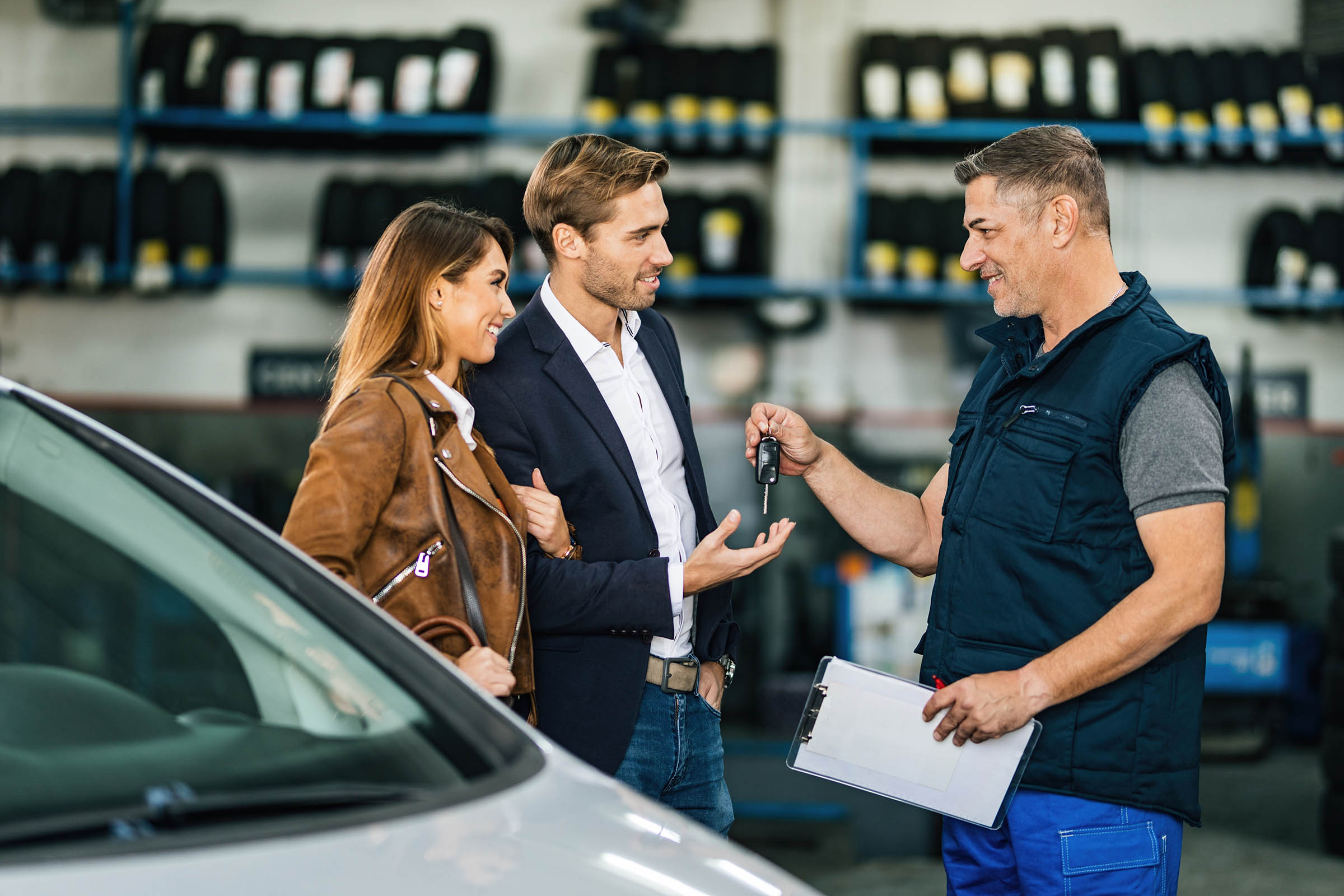 A couple receives car keys from a mechanic in a garage, surrounded by shelves of tires.