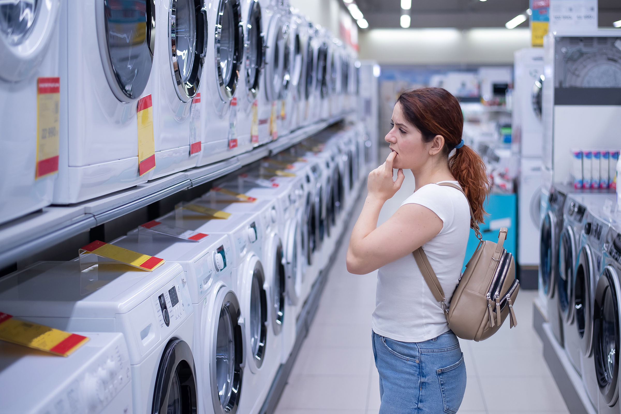 A woman with a backpack examines washing machines, appearing thoughtful in a bright, modern appliance store.
