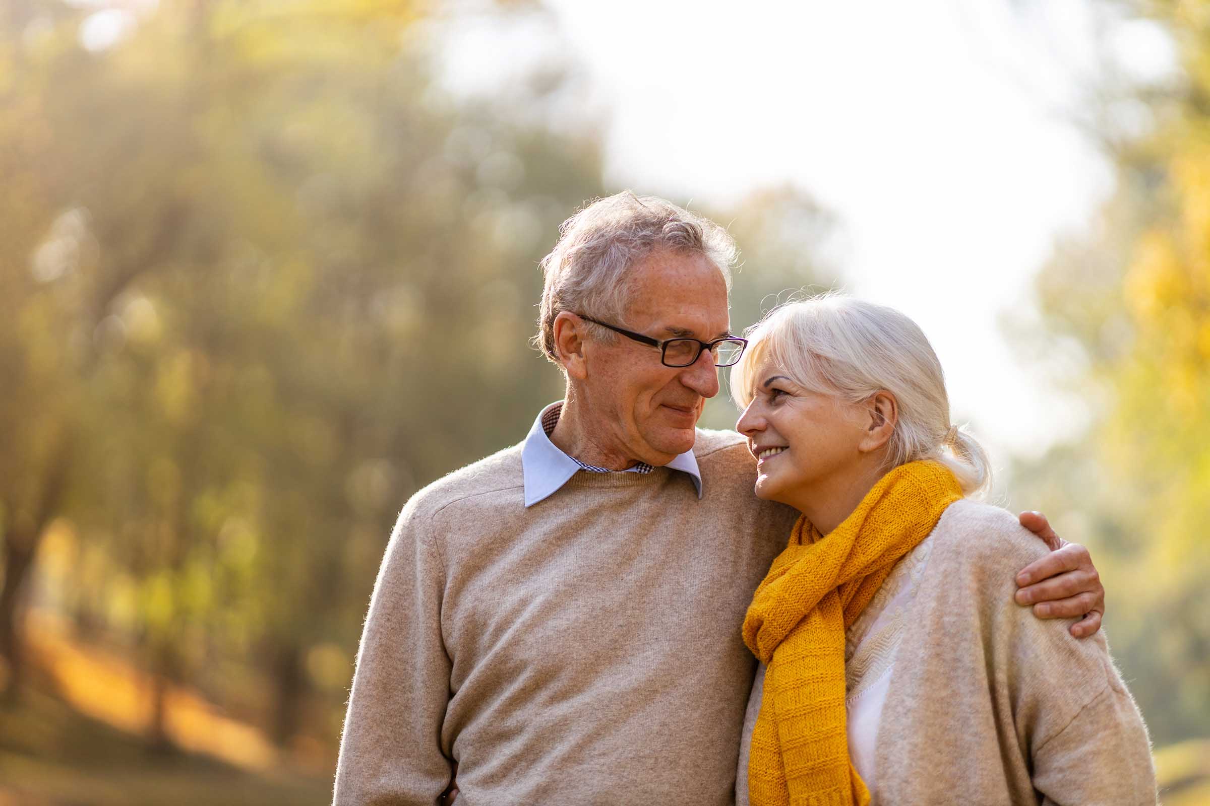 Smiling older couple in sweaters embracing outdoors, with the woman wearing a yellow scarf and trees in the background.
