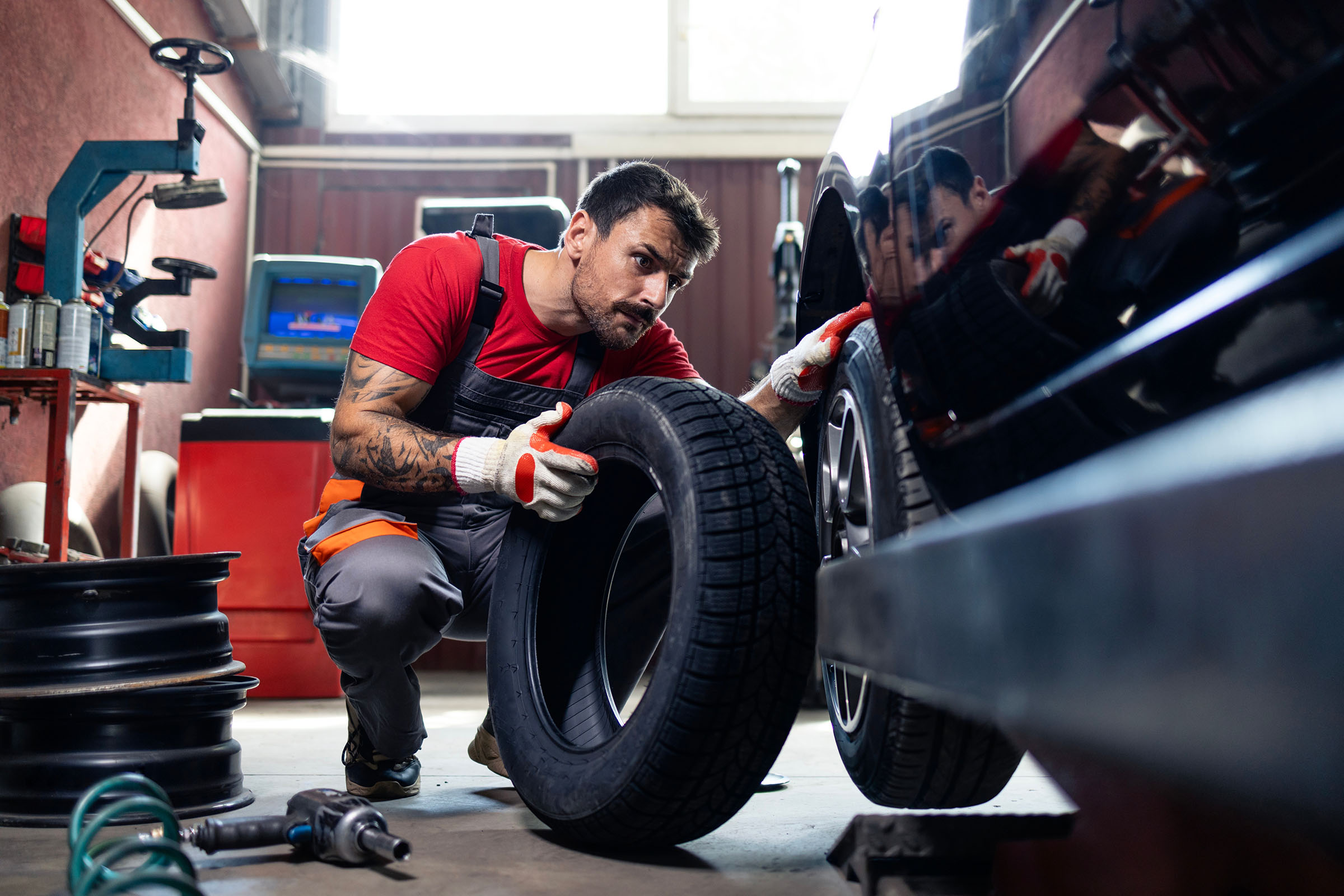 Mechanic in a red shirt and gloves replacing a car tire in a garage, holding a new tire next to the vehicle.