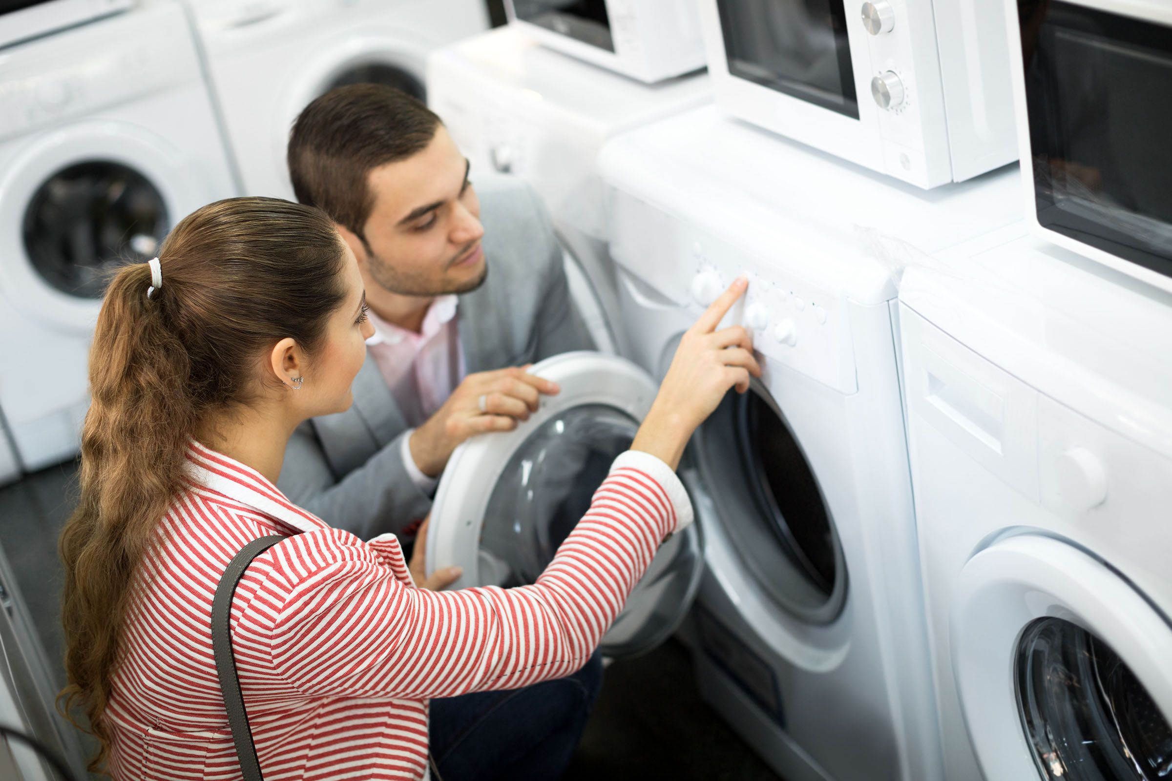 Couple examining the control panel of a front-loading washing machine in an appliance store, with microwaves stacked above the units.