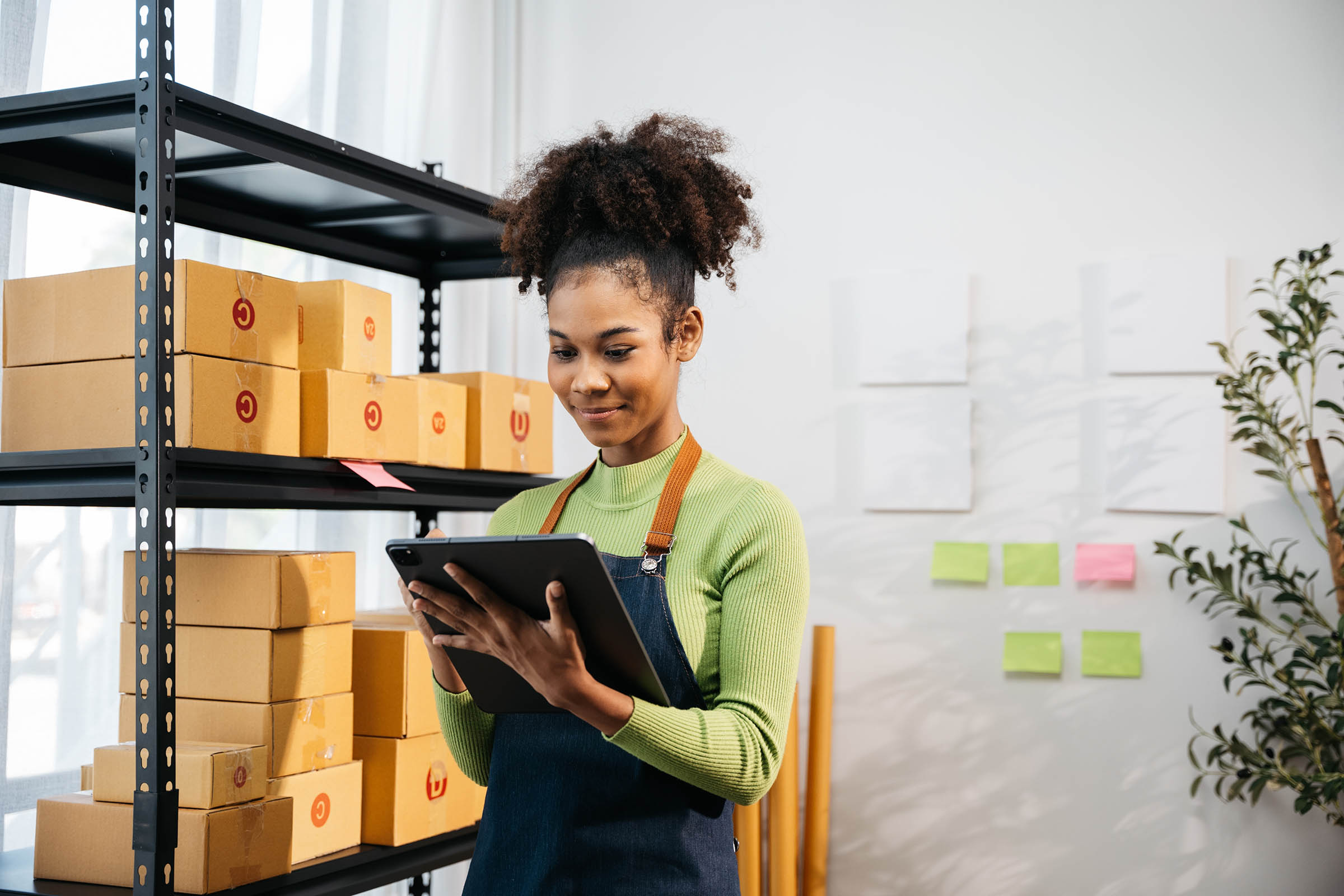 Woman with curly hair in a green sweater and apron uses a tablet in a room with shelves of boxes and sticky notes on the wall.